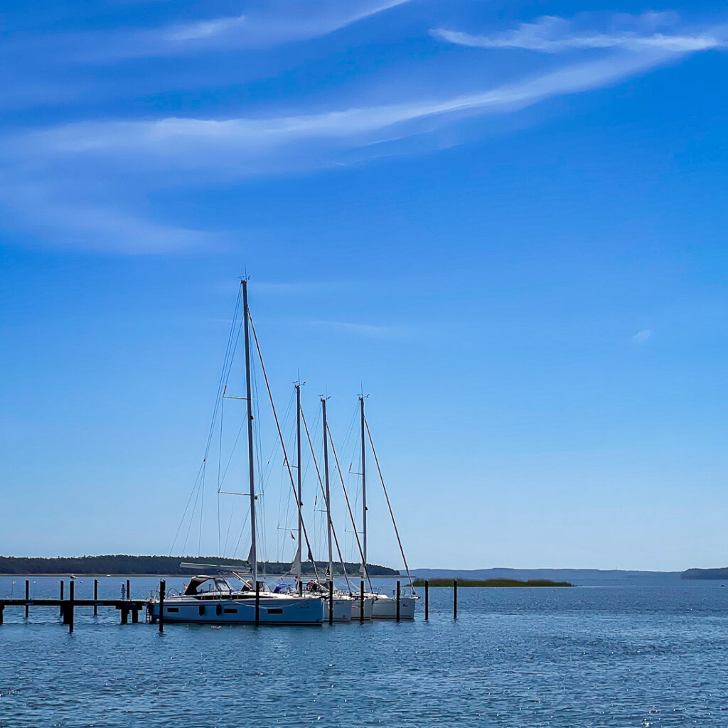 Drei Segelboote liegen an einem Steg an einem ruhigen Gewässer unter einem klaren blauen Himmel.