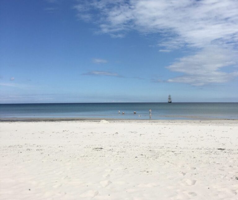 Sandstrand mit ruhigem, blauem Meer, ein paar Menschen im Wasser und ein Schiff am Horizont.