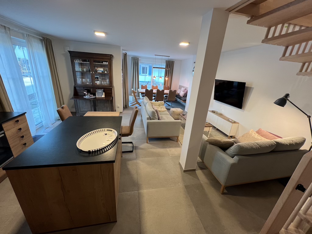 Offen gestalteter Wohnbereich mit Kücheninsel, Esstisch, Sofas, TV und großen Fenstern.