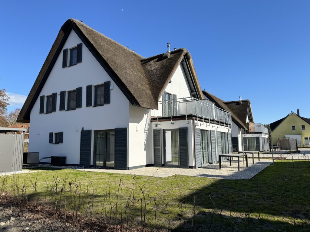 Zweistöckiges Haus mit Strohdach, weißen Wänden und schwarzen Fensterläden an einem sonnigen Tag.